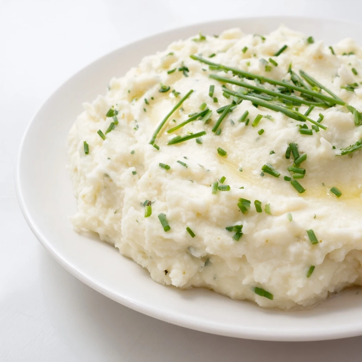 A bowl of low-carb Roasted Garlic Mashed Cauliflower topped with chopped chives, served alongside grilled chicken for dinner.